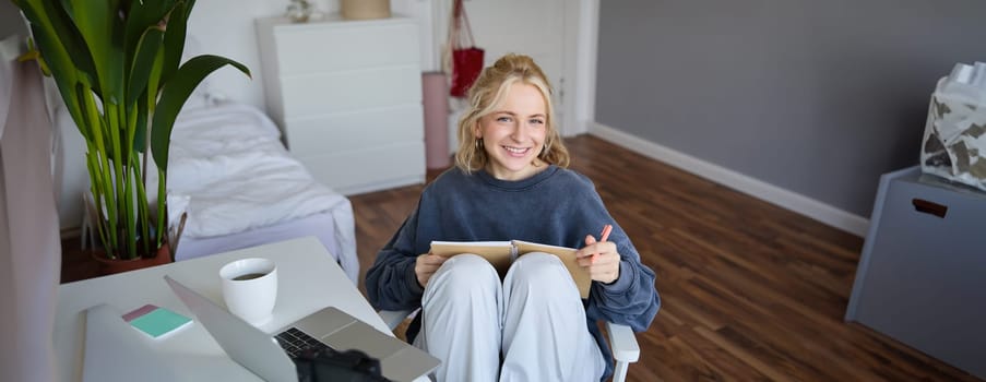 Portrait of young woman reading her notes on journal, sitting at home on chair, smiling and looking happy, doing homework.