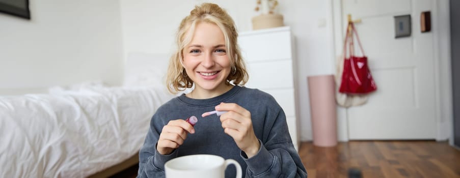 Lifestyle, beauty blogger, woman recording video of her putting on makeup, talking to camera, making online tutorial, showing her lip gloss or lipstick to followers, sitting on floor with cup of tea.