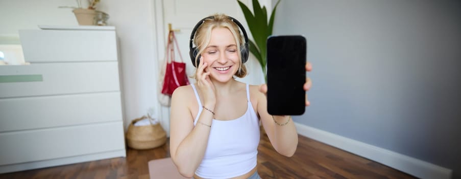 Portrait of smiling, beautiful young woman, showing her smartphone screen, wearing headphones, looking satisfied with sound quality.