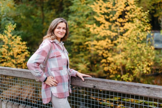 Portrait of cute young woman in casual wear in autumn, standing on bridge against background of an autumn Park and river. Pretty female walking in Park in golden fall. Copy space. smiling girl in the park standing on wooden bridge and looking at the camera in autumn season