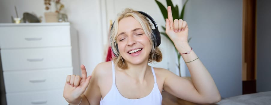 Portrait of happy and carefree blond woman, listens to music in wireless headphones, dancing and singing along favourite song, spending time alone at home.