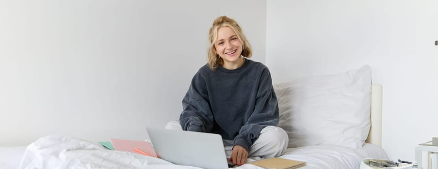 Portrait of young smiling woman studying in her bed, working from home in bedroom, sitting with laptop and notebooks on lotus pose, looking happy and relaxed.