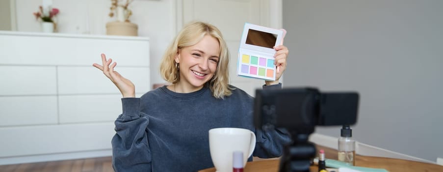 Lifestyle blogger, records video in her room, has a camera on coffee table, shows eyeshadow palette to her followers, does makeup tutorial, vlogger working indoors, creates content for social media.