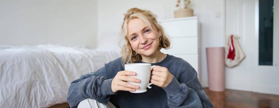 Image of young teenage girl sitting in her bedroom on floor, drinking cup of tea and enjoying day at home, smiling and looking at camera.