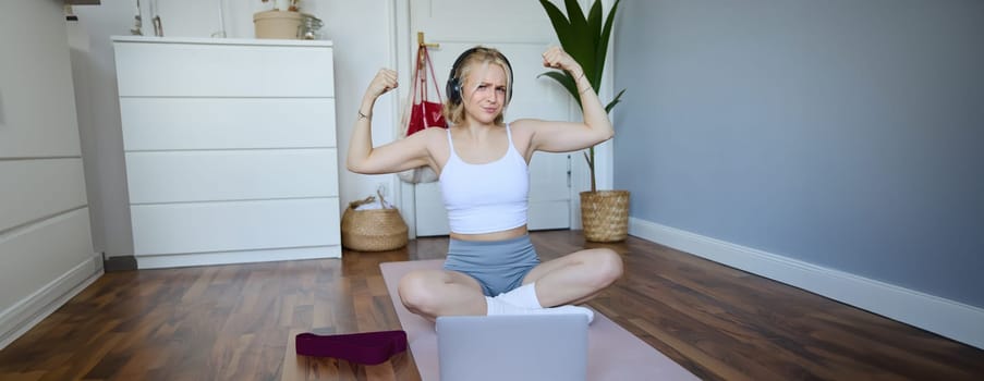 Portrait of young sporty woman following online video instructions during fitness workout, using laptop, sitting on yoga mat.