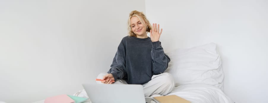Portrait of beautiful, smiling young woman sitting on bed, waves her hand at laptop camera, says hello to friend on video chat app, studies at home on remote, connects to online course.