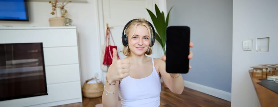 Portrait of young fitness instructor in headphones, showing smartphone screen and thumbs up, recommending app for home workout.