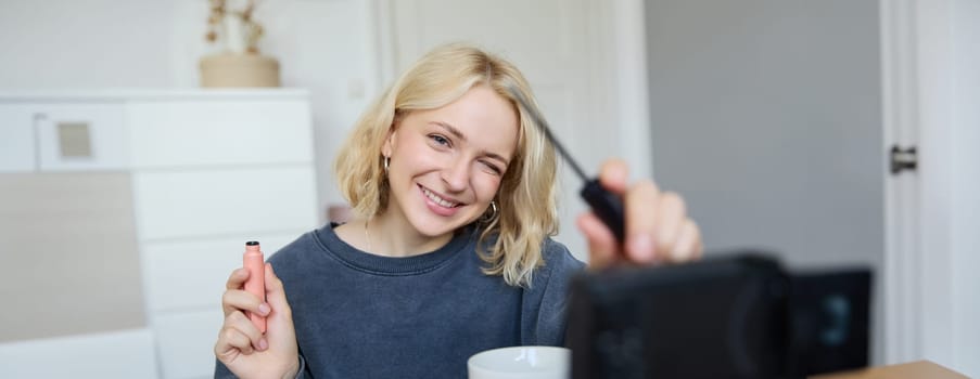 Smiling young beautiful lifestyle blogger, girl records a beauty vlog in her room, using professional video camera, showing makeup cosmetics to her followers, creates content for social media.