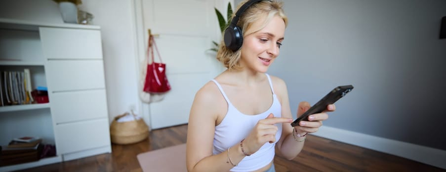 Close up portrait of athletic young woman, holding mobile phone and wearing wireless headphones, workout at home, using sport app during fitness training.