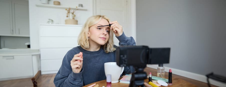 Portrait of beautiful, smiling blond woman, girl recording video of her makeup tutorial for social media, vlogger sitting on floor in her room, using stabiliser to create content, reviewing mascara.