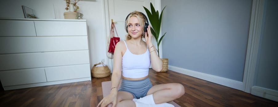 Portrait of happy, beautiful athletic woman, working out at home, listening music to boost energy while active training session, using wireless headphones and rubber yoga mat.