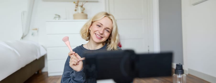 Close up portrait of happy young beauty blogger, records lifestyle vlog in her room, using camera with stabiliser, shows makeup brush and cosmetics.