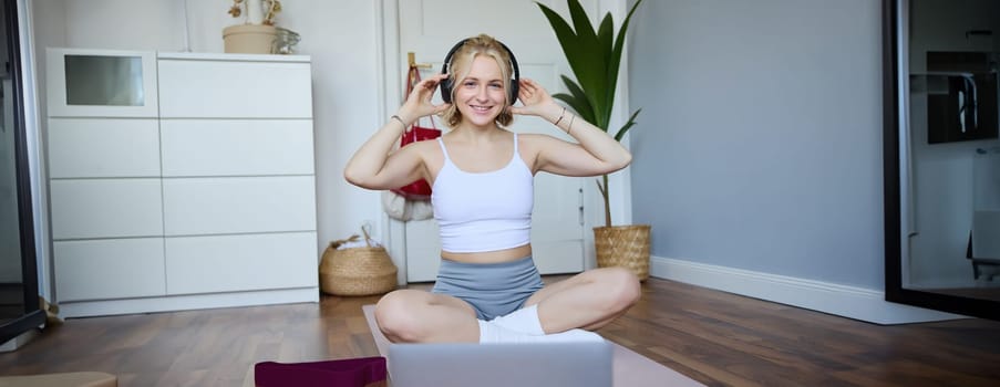 Portrait of cute young fitness woman, connects to online training session via laptop, wearing wireless headphones during workout, sits at home on rubber yoga mat.