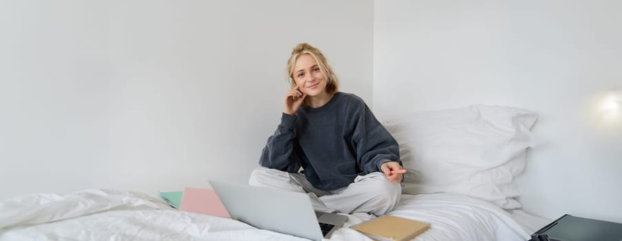 Portrait of young woman working from home, sitting on a bed with notebooks, documents and laptop, looking happy and smiling at camera.