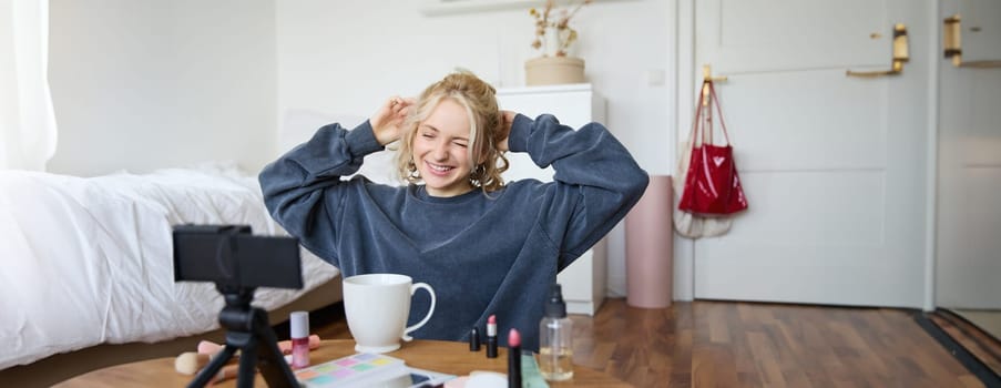 Portrait of cute smiling woman, blond girl drinks tea and records a casual, lifestyle video blog, vlogger sits in a room with camera and stabiliser, holds cup of coffee and talks.