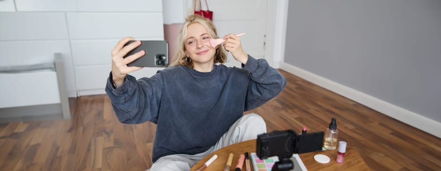 Portrait of beautiful young lifestyle blogger, woman vlogger recording video on digital camera, live stream makeup tutorial on smartphone app, sitting in bedroom, using brush on her cheeks.