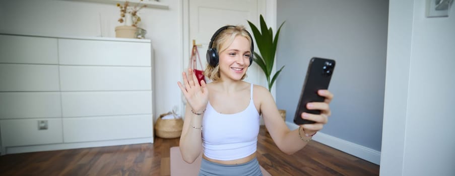 Portrait of young woman with smartphone and headphones, waving hand at mobile phone camera, live streaming during workout. Wellbeing and sport concept