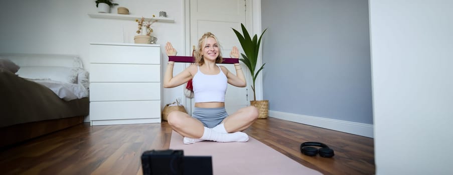 Portrait of young blond woman, social media vlogger using digital camera during workout, shooting video about exercises and fitness, stretching with rubber resistance band.