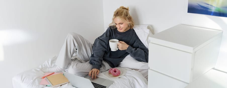 Portrait of young woman, student studying in her bed, relaxing while preparing homework, eating doughnut, using laptop in bedroom and drinking tea.