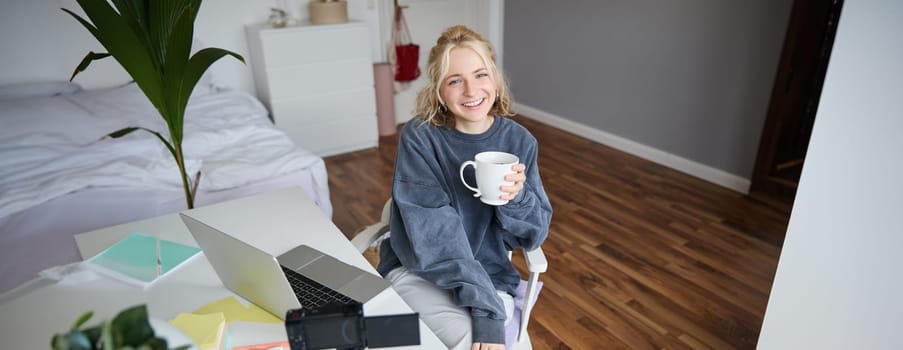 Portrait of young smiling woman, vlogger recording lifestyle video in her room, sits in front of laptop and digital camera, drinks tea, creates content for social media account.