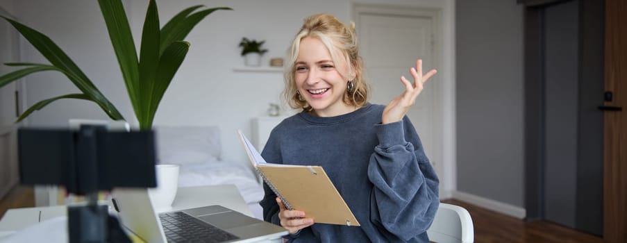 Portrait of young smiling blond woman, working from home, online chatting, using digital video camera, recording vlog, holding notebook, reading notes, explaining something.