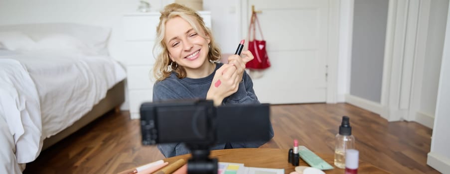 Portrait of young creative social media content creator, woman showing lipstick swatches on her hand, recording video about beauty and makeup, sitting in her room in front of digital camera.