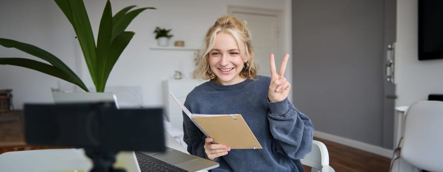 Image of smiling girl records video of herself on digital camera, shows peace sign, sits in front of laptop, holds notebook.