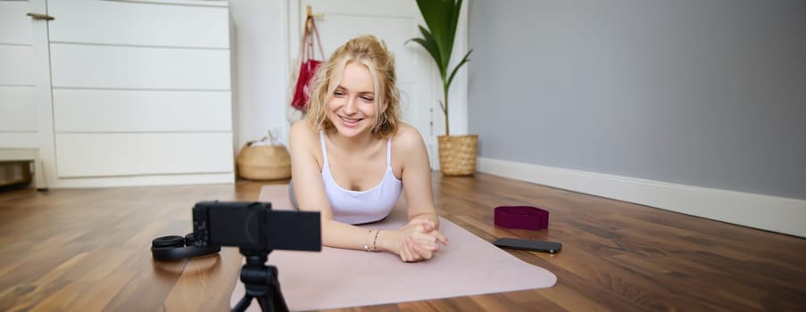 Portrait of young woman, sports vlogger, fitness instructor recording video of herself showing workout exercises, using digital camera, lying on yoga rubber mat.