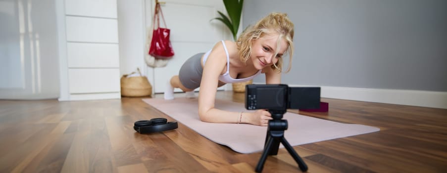 Young woman, personal fitness instructor records video of herself standing in a plank, using rubber yoga mat and digital camera at home.