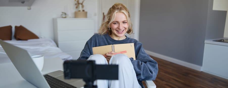 Portrait of cute, smiling young social media content creator, girl records video on digital camera and stabiliser, holds notebook, talks to audience, vlogging in her room.
