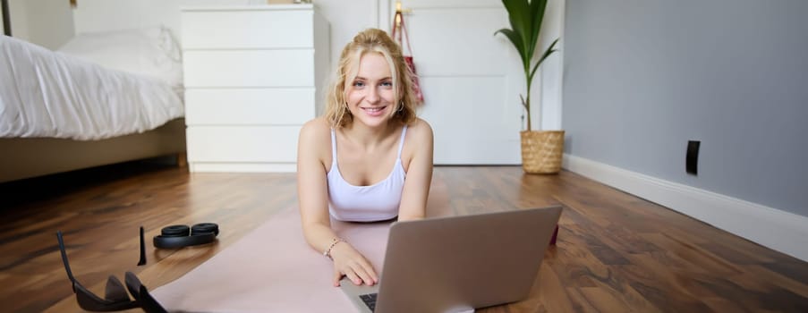 Portrait of beautiful blond woman looking at fitness video tutorials on laptop, lying on rubber yoga mat, following workout instructions online.