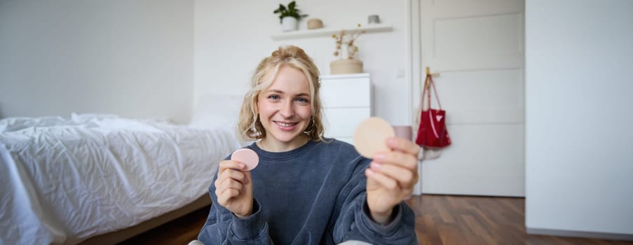 Image of smiling blond woman, vlogger, showing makeup products, beauty items, recommending to followers, sitting on floor in bedroom.