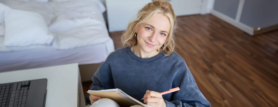 Close up portrait of beautiful young woman, writing in her diary, journal or planner, sitting in a room, smiling and looking at camera.