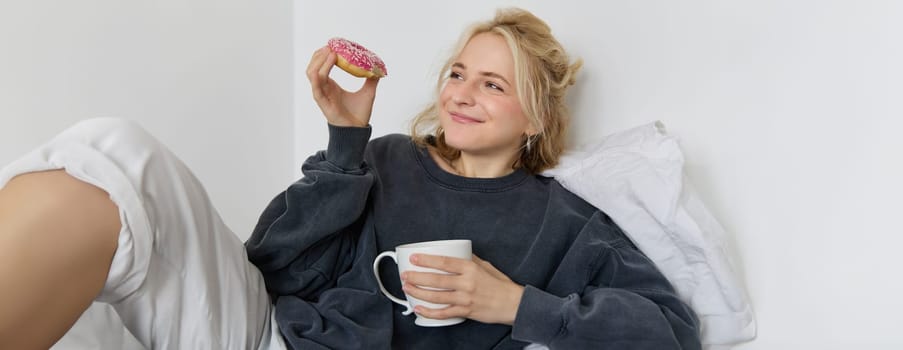 Close up portrait of beautiful smiling, blond woman, lying in bed, drinks tea and eats doughnut on a lazy weekend or day-off.