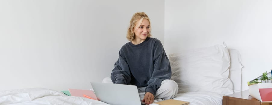 Portrait of happy blond woman, freelancer working from home, sitting on bed with laptop and notebooks. Student doing homework in bedroom, connects to online class via video chat.