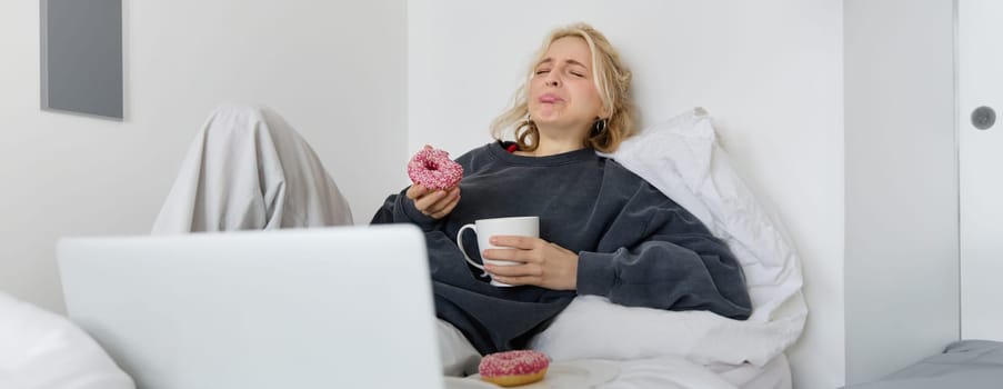 Portrait of sad and depressed woman, feeling heartbroken, lying in bed with comfort food, eating doughnut, crying from upsetting movie, watching something on laptop.