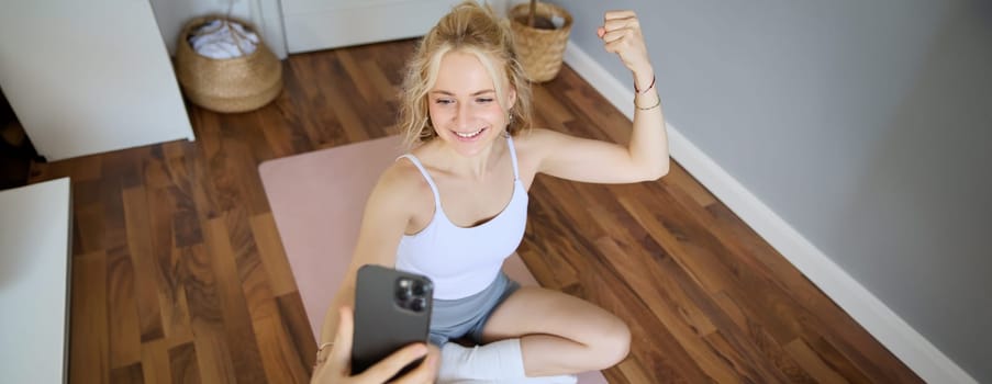 Portrait of young and fit fitness woman, training instructor sitting in a room at home, using yoga mat, flexing biceps, workout, recording herself during exercises.