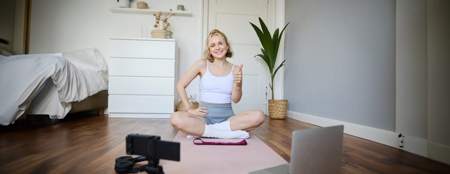 Young female athlete, fitness instructor woman sits on floor rubber mat, recording video on digital camera, showing how to workout, explaining exercises.