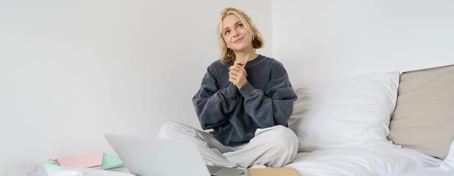 Portrait of cute smiling woman, sitting on a bed, looking dreamy, thinking, using laptop for remote work.