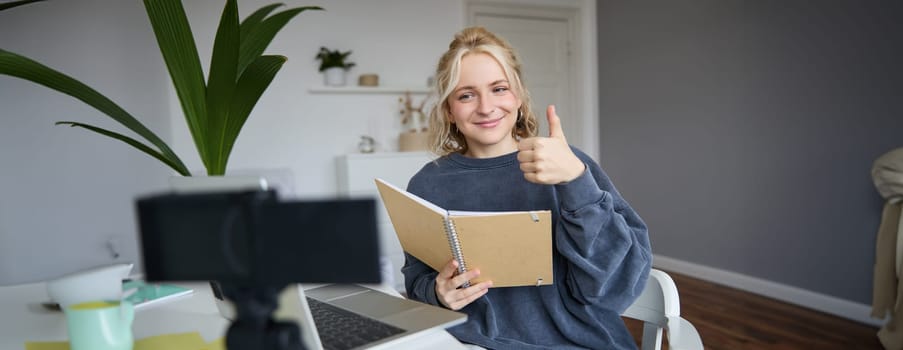 Smiling, positive young woman, shows thumbs up, sits in room with digital camera and laptop, records video, gives online tutorial, creates content for followers.