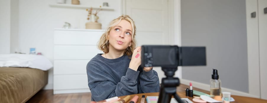 Portrait of young creative social media content creator, woman showing lipstick swatches on her hand, recording video about beauty and makeup, sitting in her room in front of digital camera.