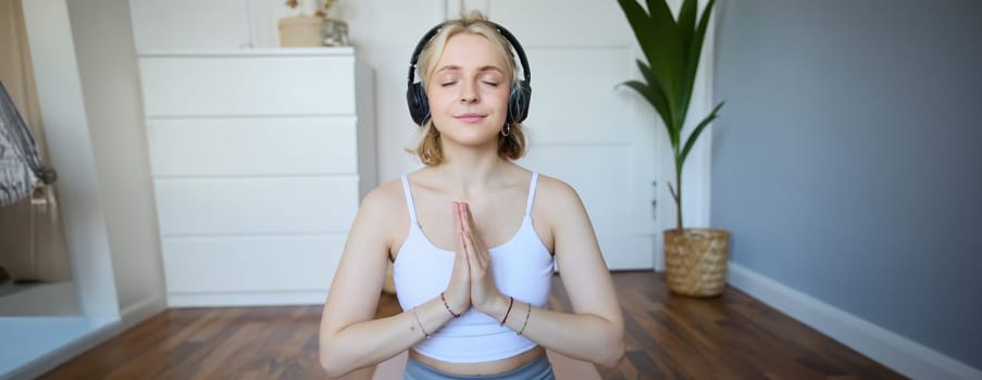 Portrait of young relaxed woman, sits in room in headphones, clasp hands together, meditated, listens to yoga music.