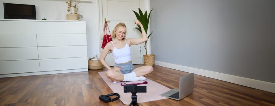 Young female athlete, fitness instructor woman sits on floor rubber mat, recording video on digital camera, showing how to workout, explaining exercises.