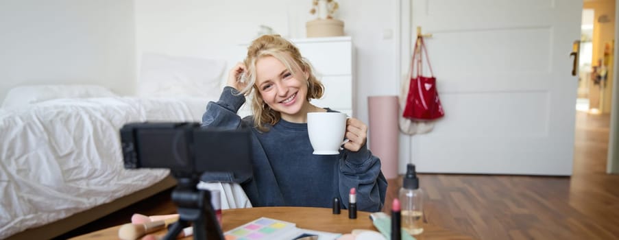Portrait of blond young lifestyle blogger, woman records video of her talking about life and beauty, sits in front of camera, holds cup, drinks tea, does blog content for social media account.