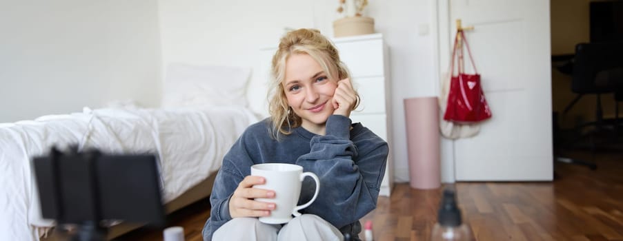 Close up portrait of beautiful blond woman, girl sits in her room, does makeup, records video for social media, drinks tea, holds mug and smiles at camera.