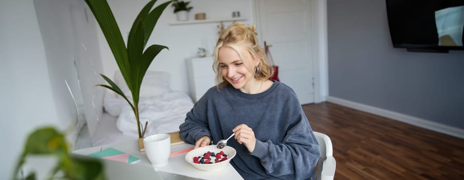 Portrait of smiling blond young woman, eating in front of laptop, watching videos online while having breakfast, enjoying dessert, sitting in bedroom.