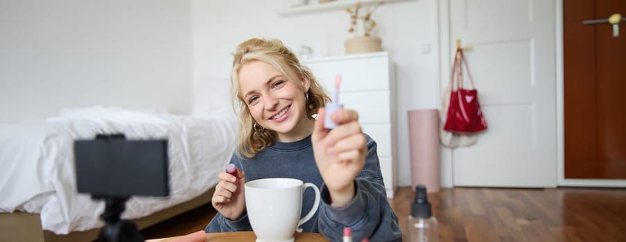 Portrait of smiling woman shows lip gloss, advertising makeup on her social media account, recording video on digital camera, content maker working on new project in her room.