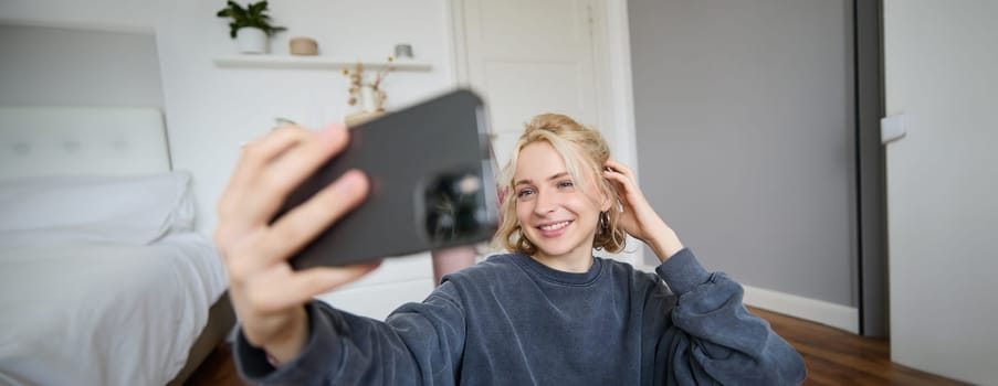 Portrait of young woman, social media influencer, taking selfies in her room, sitting on floor, holding smartphone and posing for a photo.