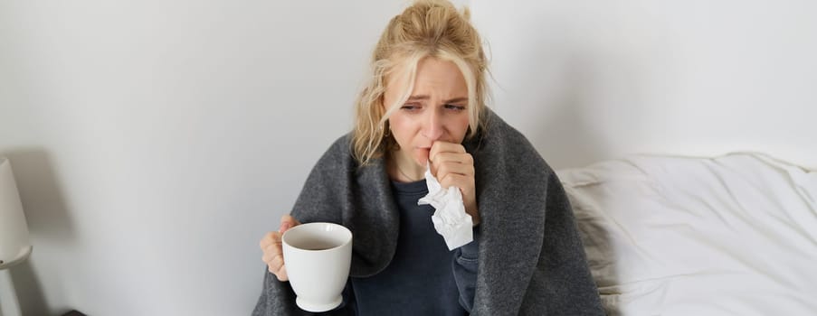 Portrait of woman catching a cold, staying home sick, drinking tea, sneezing in napkin, blowing nose, has covid symptoms.