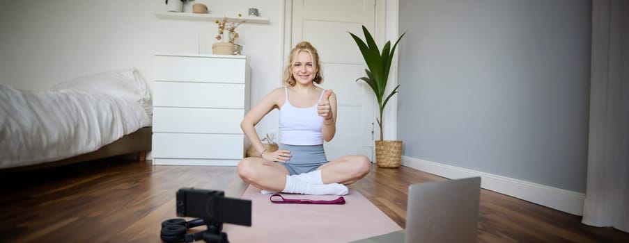 Fitness blogger, woman records video of herself, sitting on rubber mat for exercises in front of digital camera and laptop, shows thumbs up, creates content for social media account about wellbeing.